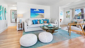 Een moderne woonkamer met een bank, salontafel en poefs, een schilderij van een strand en surfplanken aan de muur.