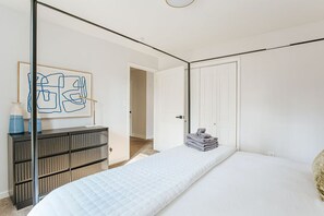 Ein Schlafzimmer mit einem Bett, einem Schminktisch, einem gerahmten abstrakten Kunstwerk und einer Tür zu einem anderen Raum.
