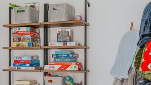 Una estantería con varios juegos de mesa y rompecabezas, un juguete de dinosaurio verde y una silla con una manta.