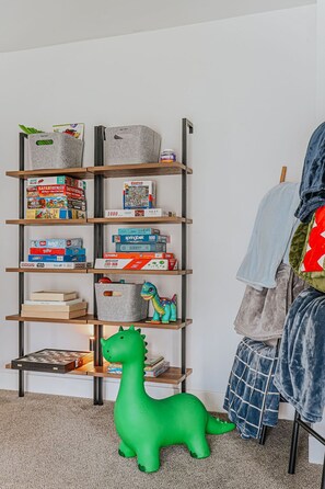 Ein Bücherregal mit verschiedenen Gesellschaftsspielen und Puzzles, eine grüne Dinosaurier-Spielzeugfigur und ein Stuhl mit einer Decke.