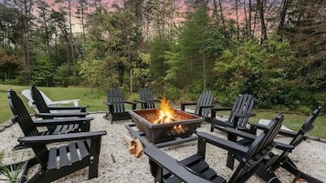 A fire pit surrounded by black Adirondack chairs in a forested outdoor area.