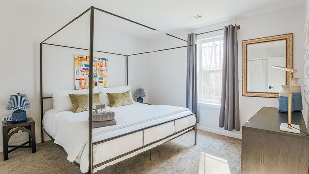 Un lit à baldaquin avec un couvre-lit et des oreillers blancs, une petite table de chevet avec une lampe, un grand miroir et une fenêtre avec des rideaux.