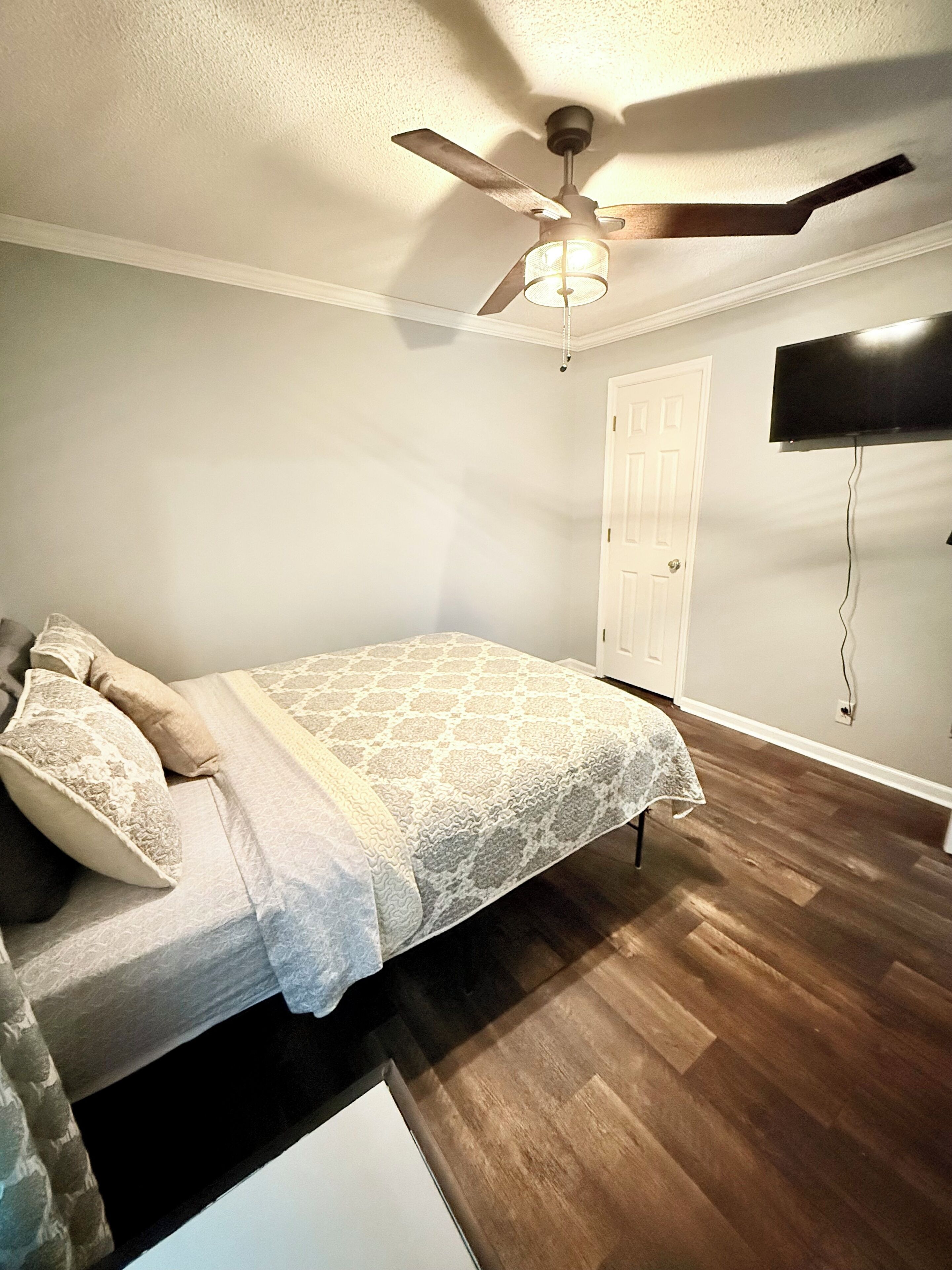 A bedroom with a bed, a ceiling fan, a TV mounted on the wall, and a door.