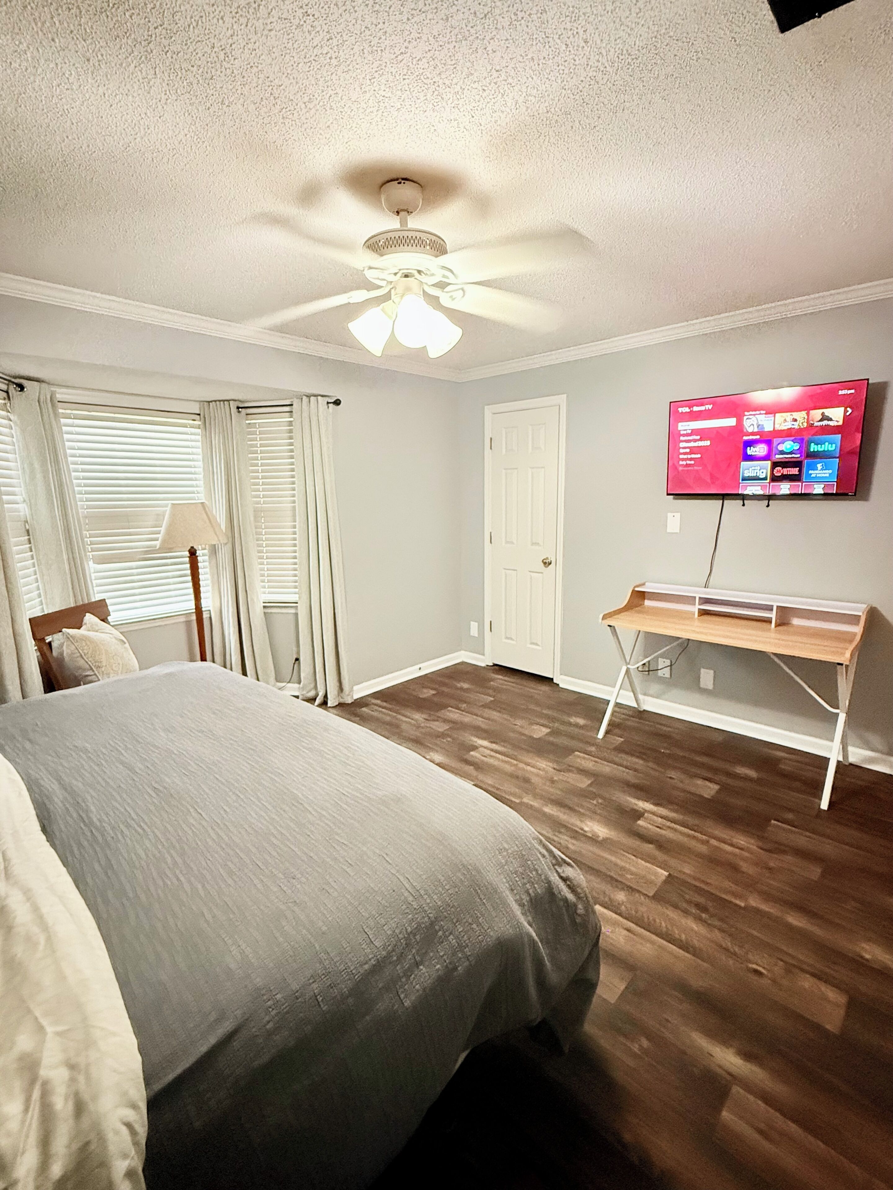 A bedroom with a bed, a desk, a ceiling fan, a TV, and a door.