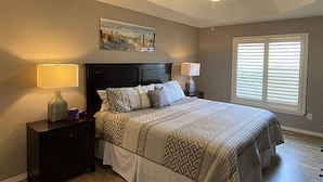 Une chambre à coucher comprenant un lit, deux tables de chevet, un ventilateur de plafond et une fenêtre avec des stores.