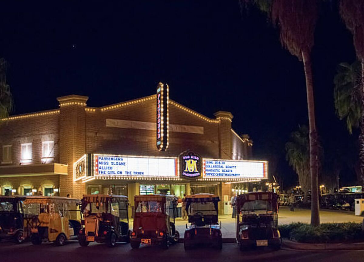 A theater with a marquee displaying movie titles and a row of vintage cars parked in front.
