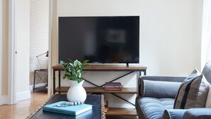 Ein Wohnzimmer mit Flachbildfernseher auf Holzfuß, Couchtisch mit Buch und Pflanze, sowie ein graues Sofa.
