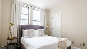 A neatly arranged bedroom with a large bed, two bedside lamps, a window with curtains, and a door. - Downtown Boston - close to everything - Laundry in Building, Keyless Entry. Free Air Mattress too. (Boston)