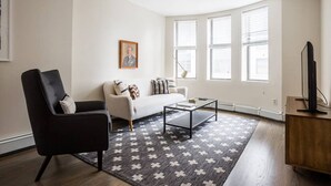 A living room with a black armchair, a white sofa, a glass coffee table, and a patterned rug. There are three windows with blinds, a framed picture on the wall, and a television on a wooden stand. - Downtown Boston - close to everything - Laundry in Building, Keyless Entry. Free Air Mattress too. (Boston)
