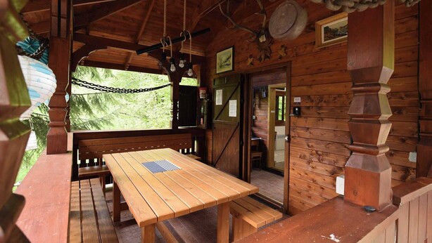 Eine Holzhütte mit Holztisch und -bänken, einer Tür, die nach draußen führt, und einer Kette an der Decke.