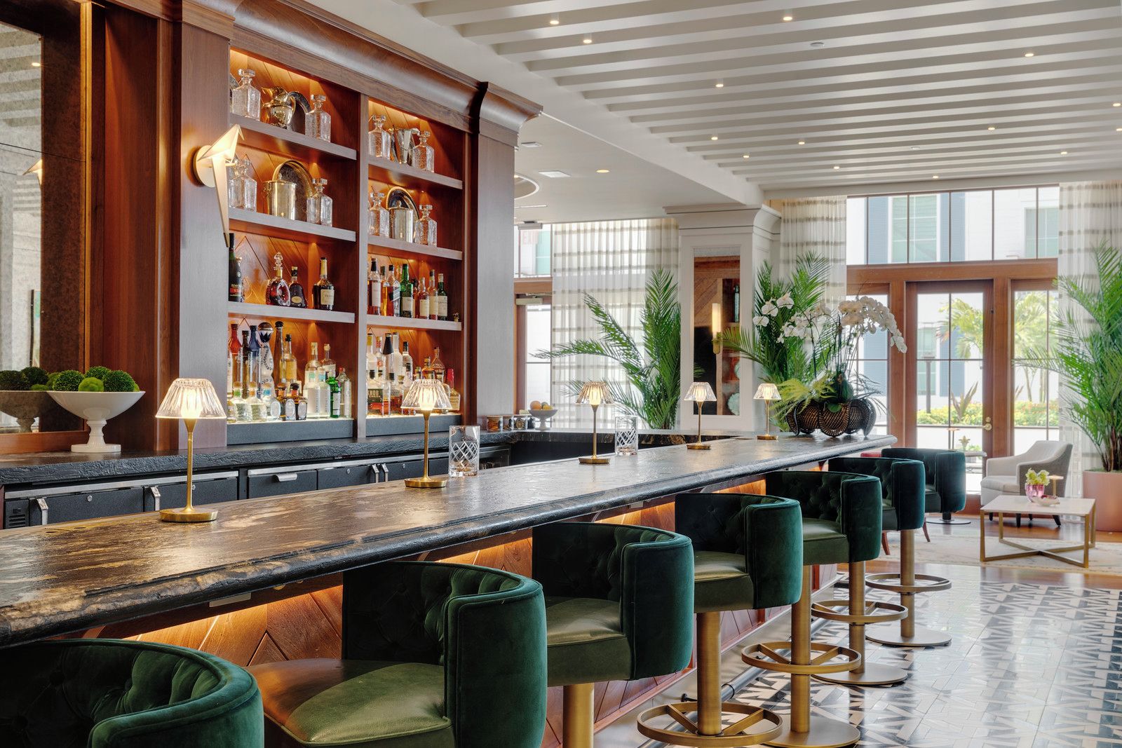 Uma área de bar com um balcão comprido, banquetas estofadas verdes e prateleiras com garrafas.