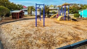 A playground with swings and slides, surrounded by a sandy area and residential buildings in the background. - In Sea State Escape (Kure Beach)