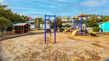A playground with swings and slides, surrounded by a sandy area and residential buildings in the background.