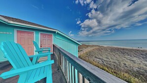 Un bâtiment turquoise avec des portes roses, une terrasse en bois et une vue sur la plage et l’océan.