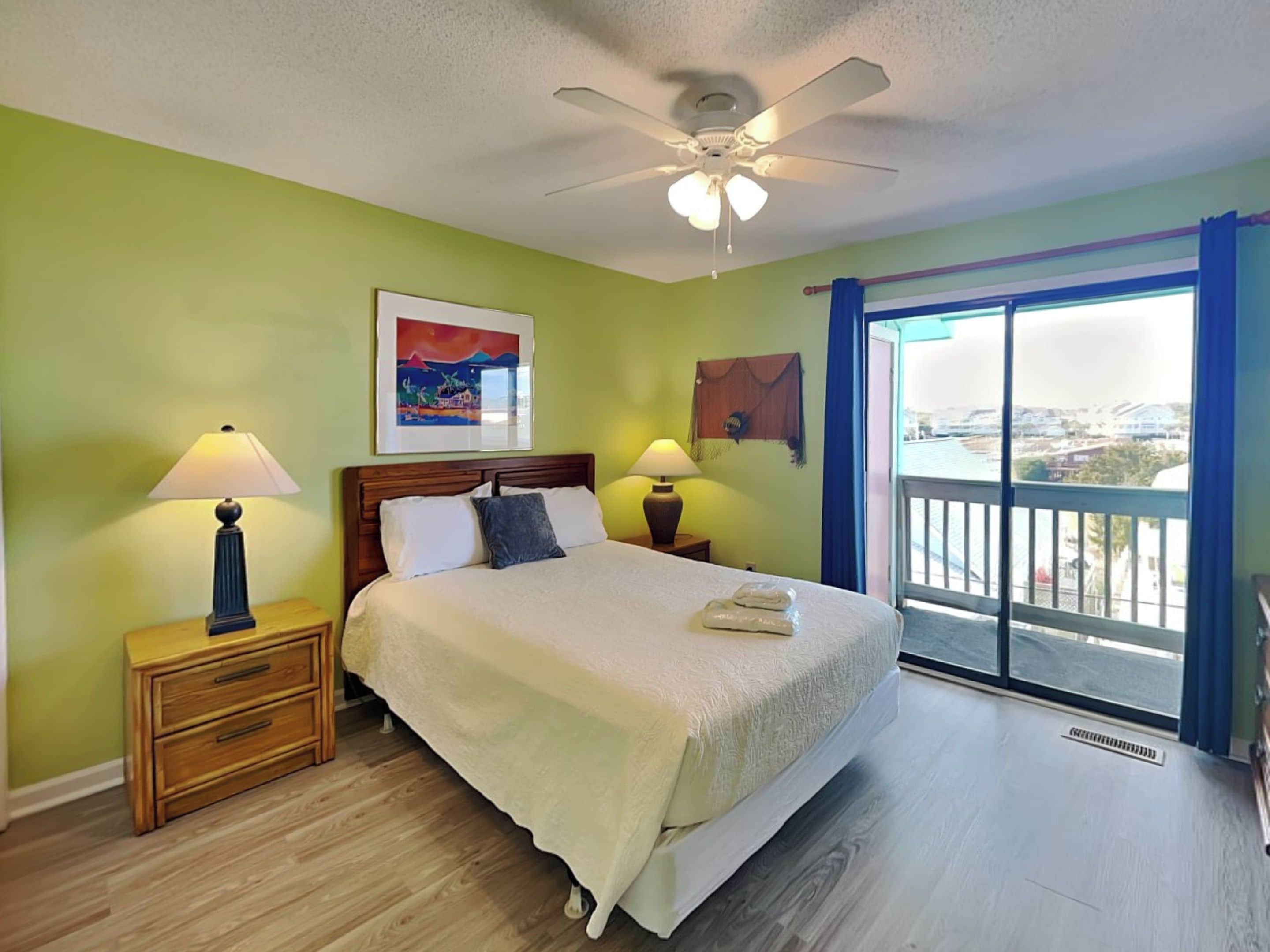 A hotel room with a bed, two bedside tables with lamps, a ceiling fan, a painting on the wall, and a view through a large window.