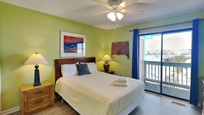 Une chambre d’hôtel avec un lit, deux tables de chevet avec des lampes, un ventilateur de plafond, un tableau au mur et une vue à travers une grande fenêtre.