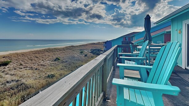 Une vue sur la plage depuis une terrasse en bois, avec des chaises turquoise et une table, donnant sur l’océan et la plage.