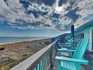 Vista de la playa desde una terraza de madera con sillas turquesas y una mesa, con vista al océano y la playa.