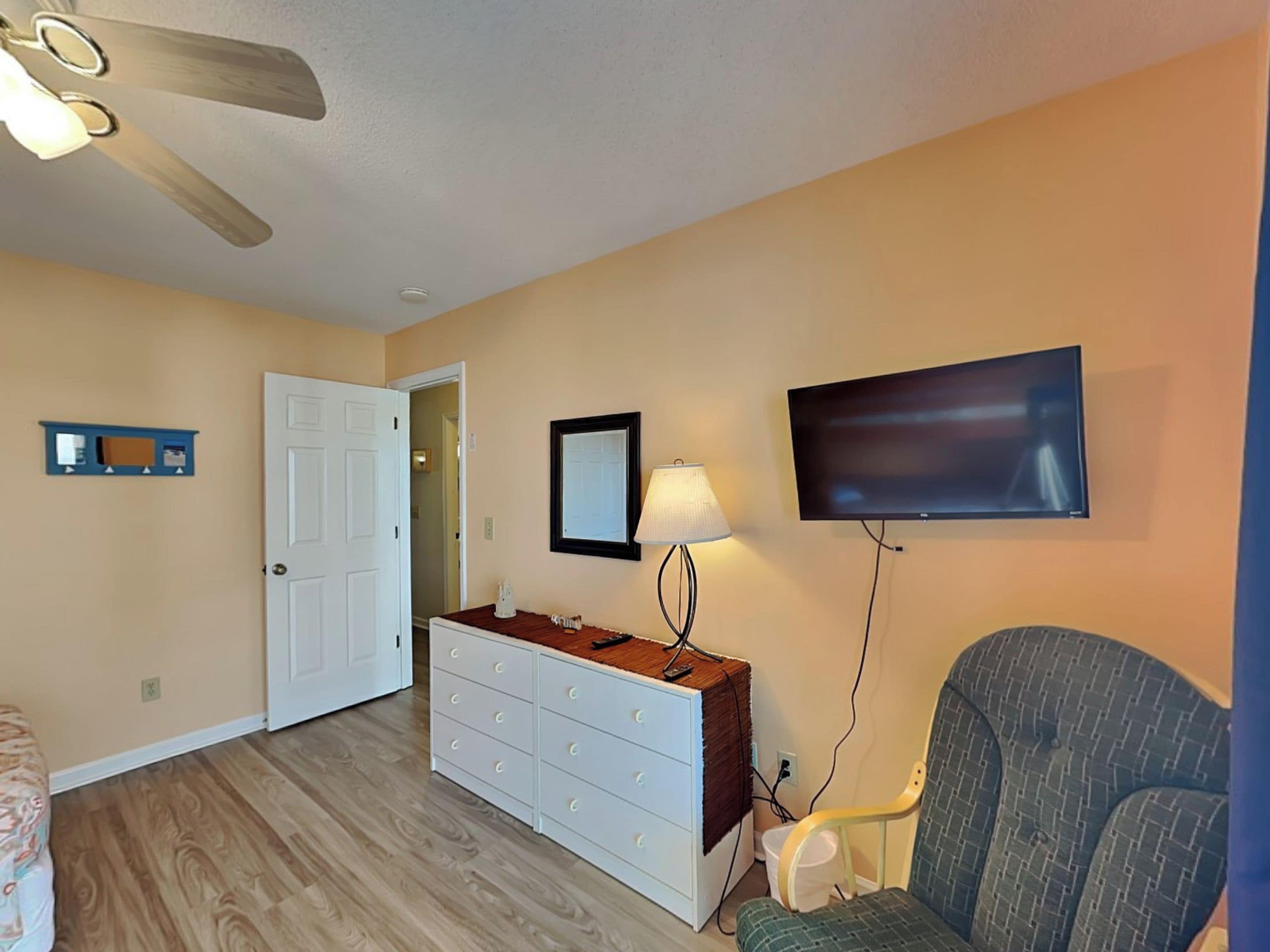 A bedroom with a ceiling fan, a dresser, a chair, a TV, and a door.