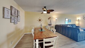 A dining area with a wooden table and white chairs, a blue sofa, and a ceiling fan. - Winds V1 #3B - My Top Paradise (Carolina Beach)