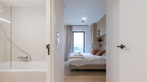 Ein modernes Schlafzimmer mit einem großen Bett, einer Duschkabine und Blick auf die Stadt durch ein großes Fenster.