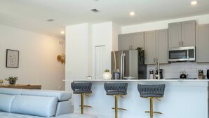 Una cucina moderna con mobili grigi, un frigorifero in acciaio inossidabile e un forno a microonde. Sono presenti tre sgabelli da bar con gambe dorate e rivestimento grigio. Un piano di lavoro bianco corre lungo la cucina, e in primo piano si intravede un divano bianco.