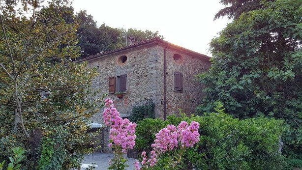 A stone building with two windows and a small balcony, surrounded by lush greenery and flowering plants. - Ai Chiosi (Pontremoli)