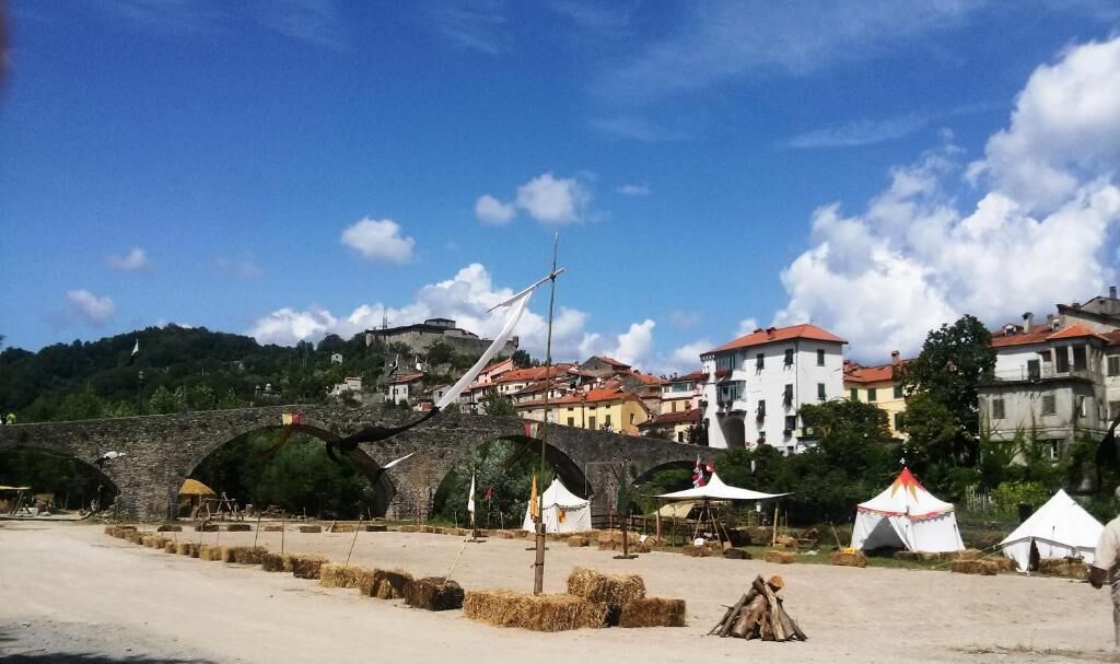 A stone bridge with arches, a sandy area with hay bales, and buildings with red roofs in the background.
