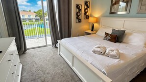 A bedroom with a bed, a nightstand, a lamp, a dresser, and a view of a pool and buildings. - 3BR Home w/ Pool Near Disney (Clermont)