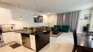 A modern kitchen with a central island, a dining area with a wooden table, and a living room with green sofas. - 3BR Home w/ Pool Near Disney (Clermont)