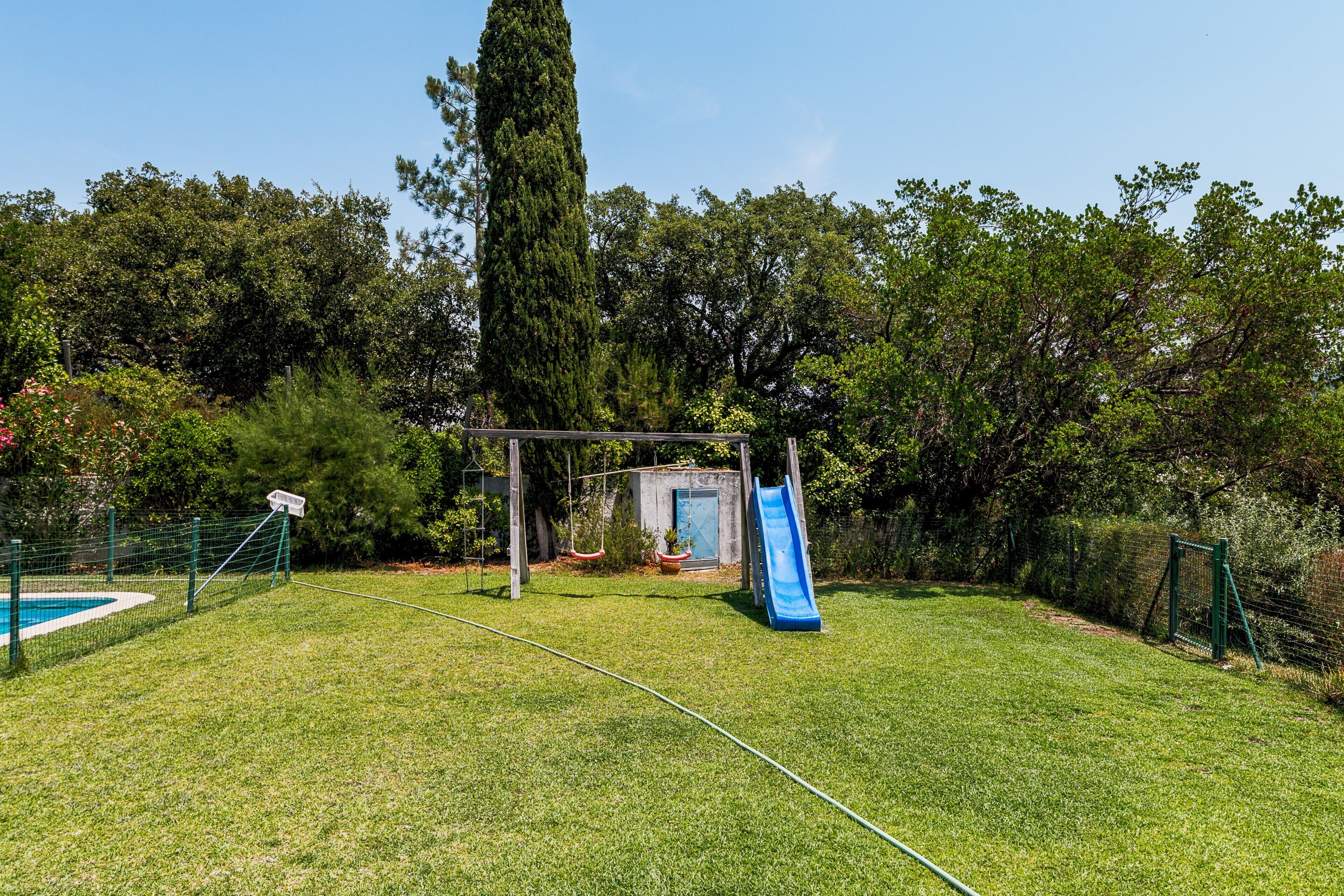 A backyard with a swing set, a slide, a fenced pool, and a tall tree.