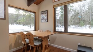 A dining area with a wooden table and chairs, large windows with a snowy view, and a framed New Yorker magazine on the wall. - Ski, Hike & Explore-Waitsfield Retreat Near Sugarbush (Waitsfield)