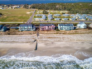 Vista aérea de una zona residencial costera con casas a lo largo de la playa, una playa de arena y olas rompiendo sobre la orilla.