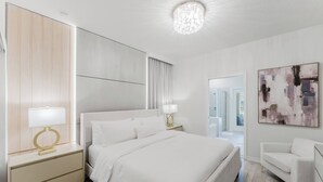 A modern hotel room with a large bed, a nightstand with a lamp, a white armchair, and a large abstract painting on the wall. - Family 7BR Villa Near Disney & Parks (Clermont)