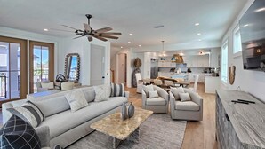A spacious living area with a sofa, coffee table, and chairs. There is a kitchen in the background with modern appliances and a dining area. - Sand & Stories: Rooftop Deck, Pet-Friendly, Sleeps 16, Sugar Sands Community (Mexico Beach)