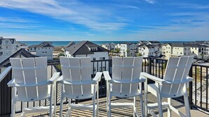A balcony with white chairs overlooking a coastal residential area with houses and the sea. - Sand & Stories: Rooftop Deck, Pet-Friendly, Sleeps 16, Sugar Sands Community (Mexico Beach)