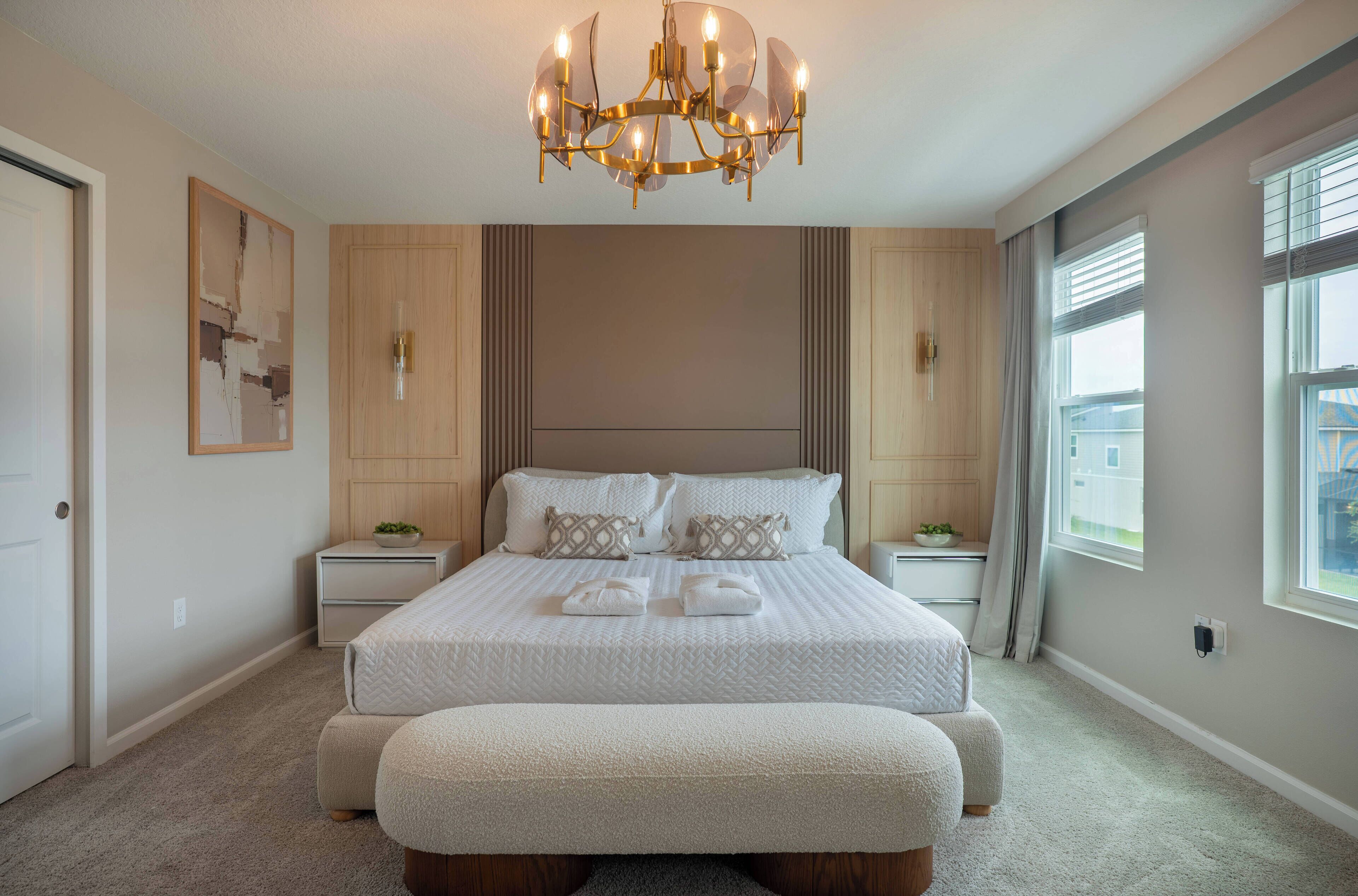 Een slaapkamer met een groot bed, een bankje, een kroonluchter en een raam met jaloezieën.