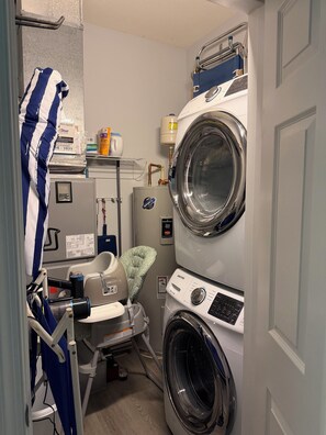 A compact laundry room with a front-loading washer and dryer, a utility cart, and a water heater. - Beach & Bay at Five Points-3 BR/2BA Pet Friendly Condo-Pool, Tennis & Pickleball (Lewes)