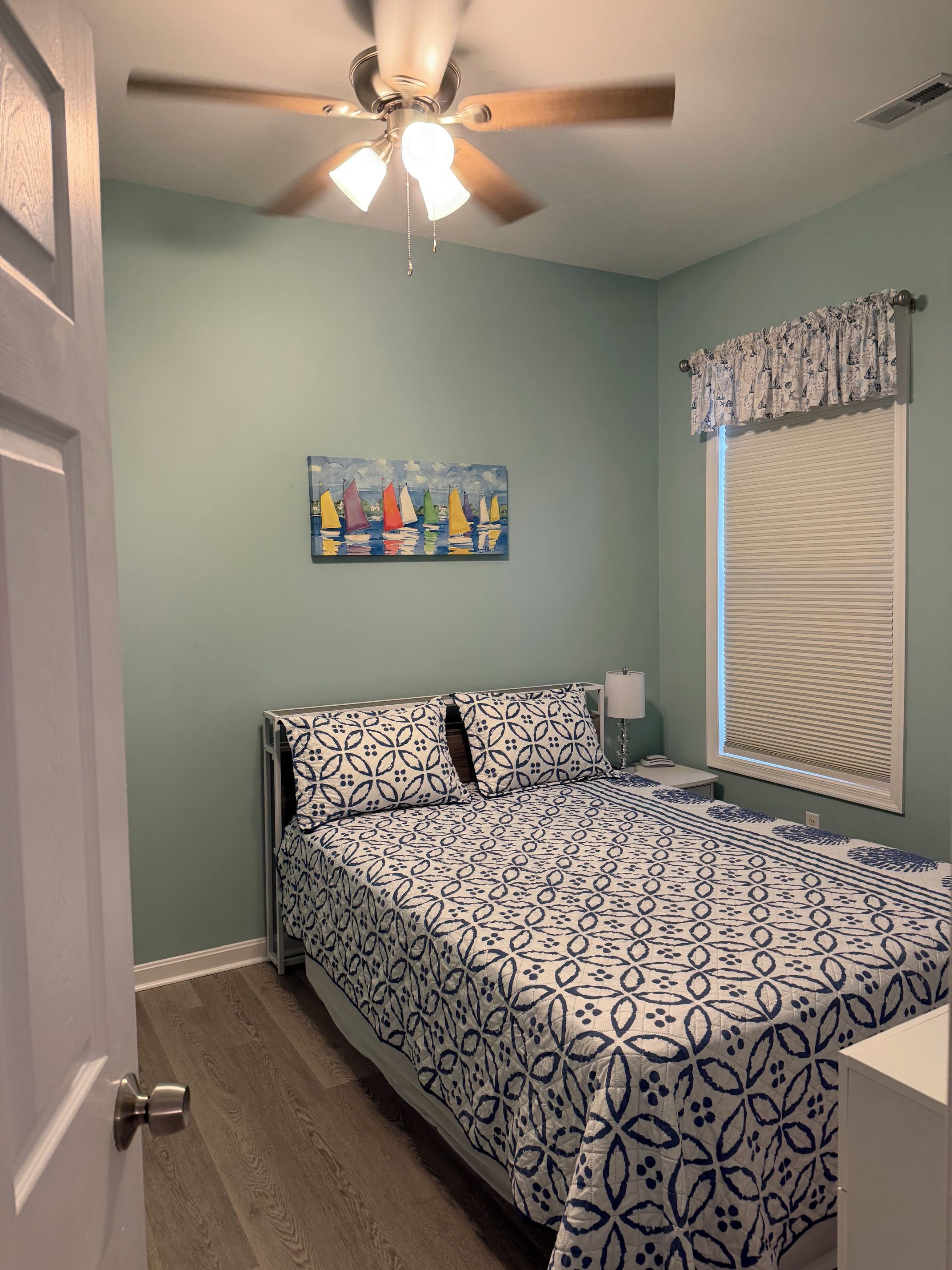 A bedroom with a bed, a ceiling fan, a painting, a window with blinds, and a door.