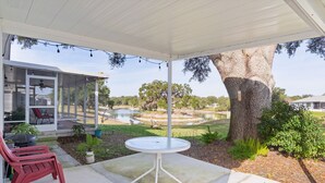 A covered patio with a table and chairs, a large tree, and a view of a pond and surrounding landscape. - Lake Paradise 2B/2B Home in Village of Orange Blossom Gardens (The Villages)