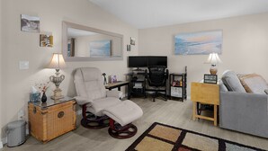 A living room with a sofa, a recliner, a TV, a desk, and a computer. There are wall art pieces and a small rug. - Lake Paradise 2B/2B Home in Village of Orange Blossom Gardens (The Villages)