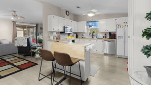 A modern kitchen with white cabinets, a central island with bar stools, a microwave, and a refrigerator with magnets. - Lake Paradise 2B/2B Home in Village of Orange Blossom Gardens (The Villages)