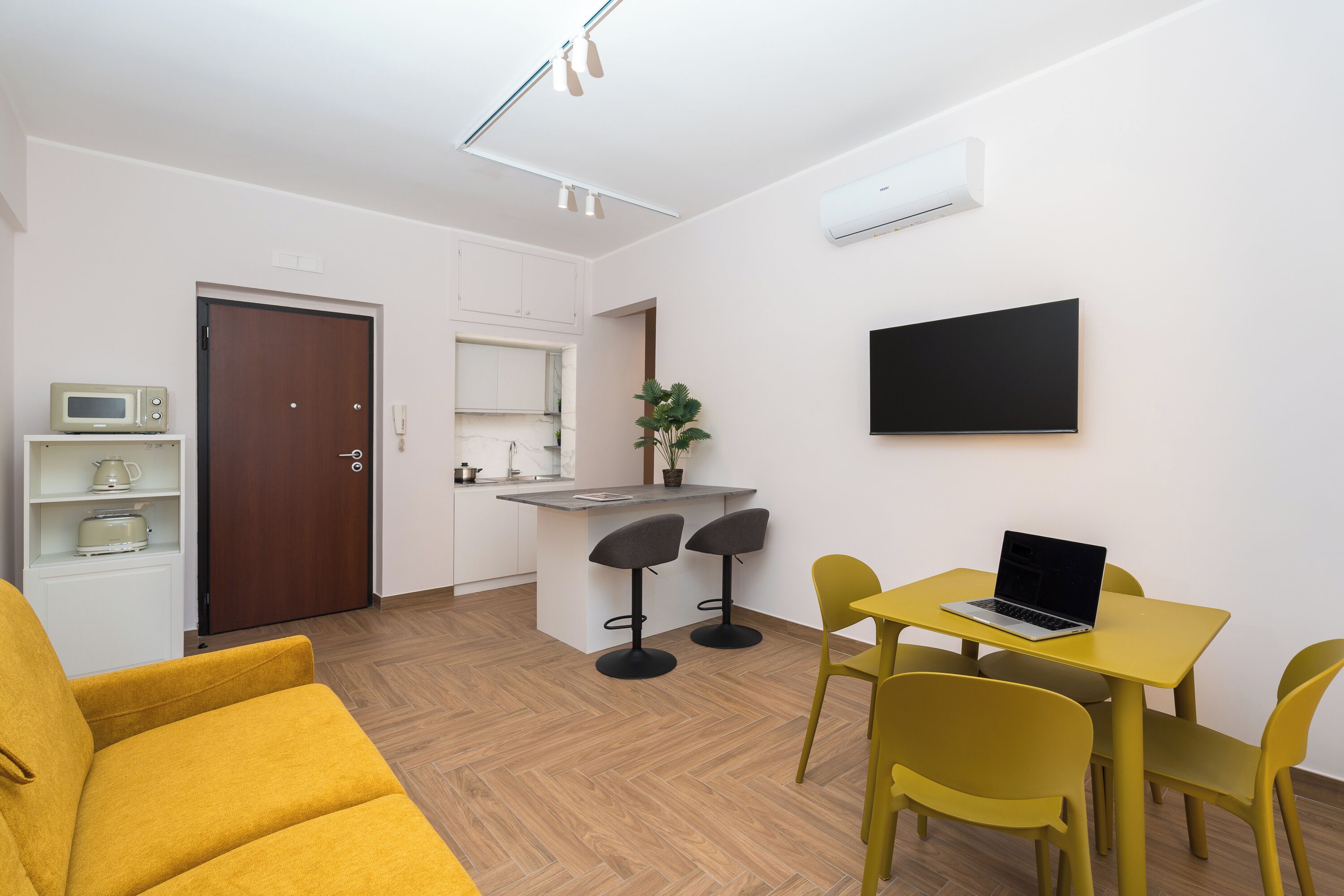 Een moderne woonkamer met een gele bank, een eettafel met gele stoelen, een keuken met een magnetron en een televisie aan de muur.