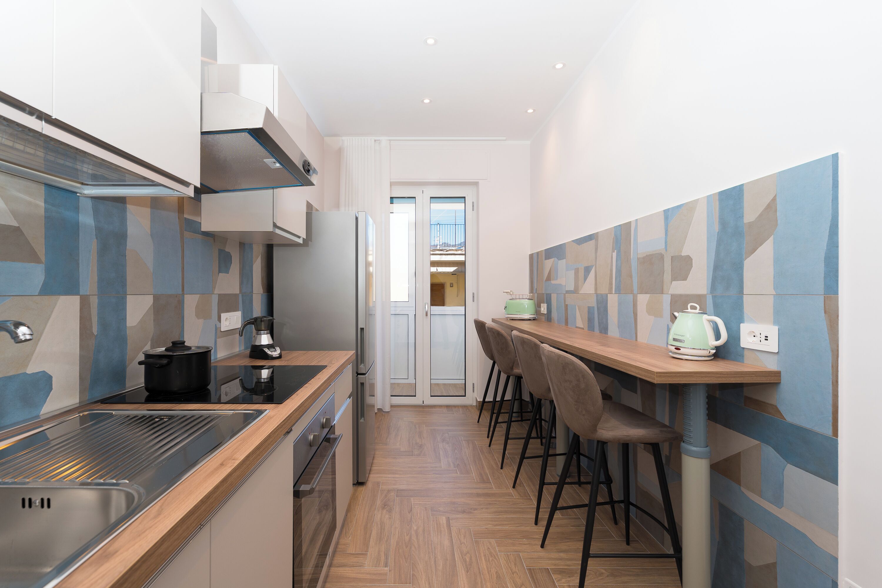 A narrow kitchen with a wooden countertop, a sink, and a stove. There is a dining area with a table and chairs.