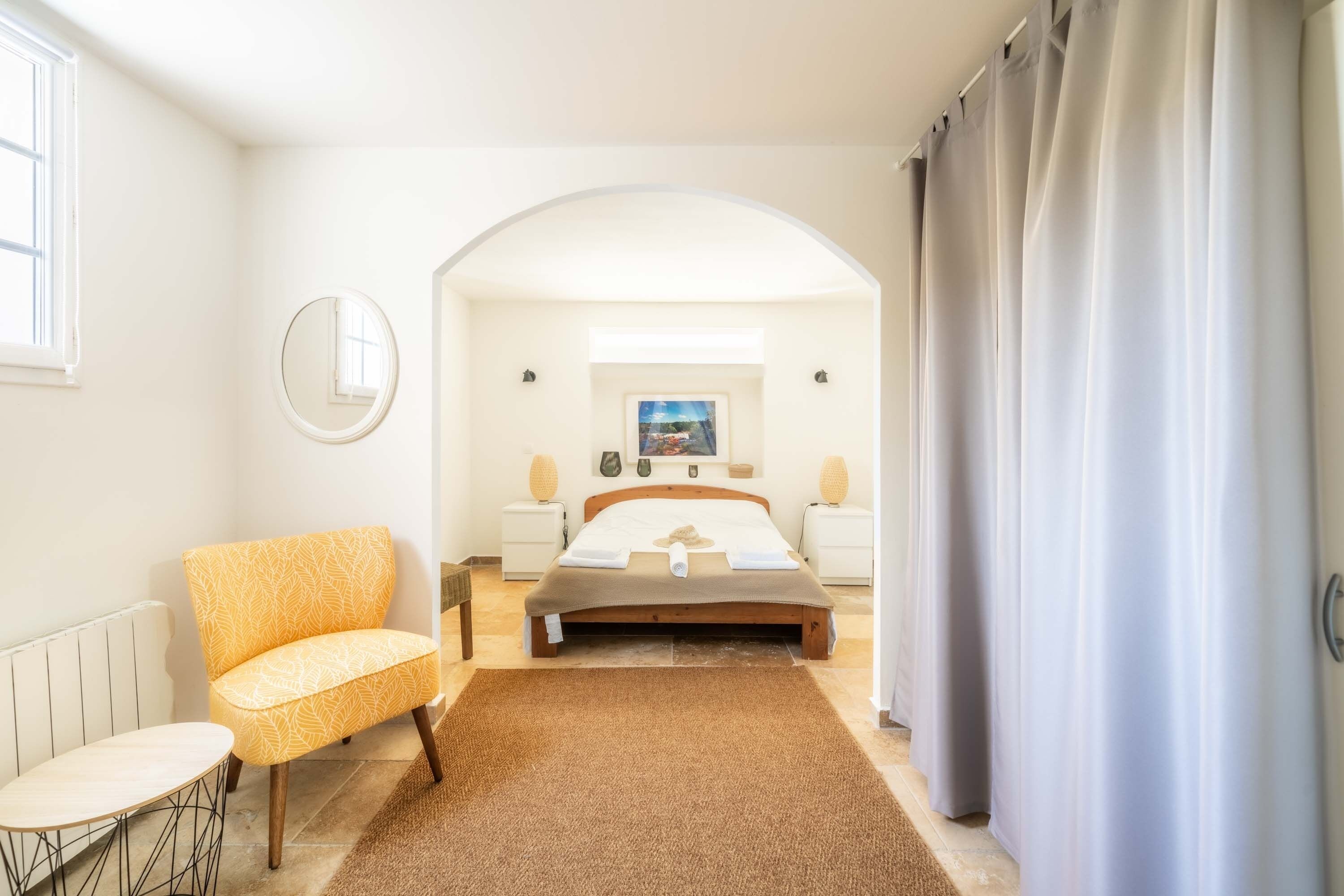 A bedroom with a bed, a chair, a small table, a mirror, and a picture on the wall.