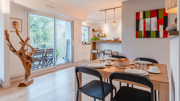 A modern dining area with a wooden table set for two, black chairs, and a view through large sliding glass doors to an outdoor patio. - Fleur de Sel - Centre de Nantes avec parking (Nantes)
