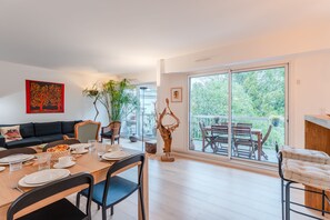 A modern dining area with a wooden table set for four, a black sofa, and a large window with a view of greenery. - Fleur de Sel - Centre de Nantes avec parking (Nantes)