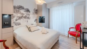 A modern hotel room with a large bed, a red chair, a desk with a lamp, and a wall-mounted TV. - Fleur de Sel - Centre de Nantes avec parking (Nantes)