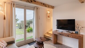 A modern living room with a large TV, wooden beams, a sliding glass door to a garden, and a cozy seating area. - La Croisée des Sentiers - Aperçu Vue Mer (Pléneuf-Val-André)
