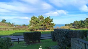 A view from a covered porch overlooking a grassy area with a stone wall and a hedge, a road, and a body of water in the distance. - La Croisée des Sentiers - Aperçu Vue Mer (Pléneuf-Val-André)